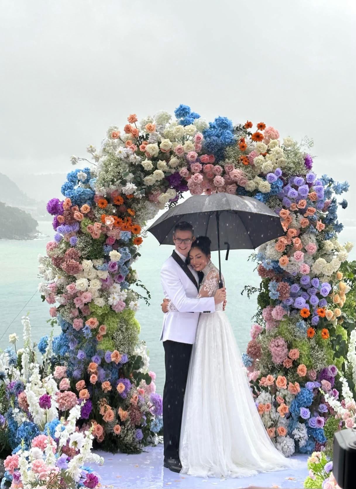 Couple under an umbrella beneath a flower arch above the sea at a villa wedding at Samsara, Kamala, Phuket